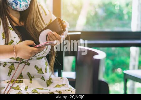 Asiatische weibliche Kunden tragen Gesichtsmaske mit Smartphone, während sitzen in der Warteschleife soziale Distanz für Take-Away-Drink im Café. Neue normale Lebensdauer Stockfoto