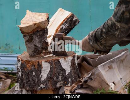 Ein Baumstamm mit einer Axt auf einem Deck mit männlichen Händen in Stoffhandschuhen und einer Tarnjacke im Hinterhof in die Hälfte gespalten. Hausarbeit zur Vorbereitung auf das wi Stockfoto