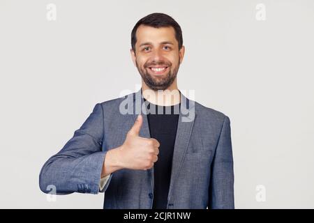 Junger Geschäftsmann mit Bart in einer Jacke, mit einem glücklichen Lächeln, zeigt seinen Daumen zur Kamera. Alles ist gut, es zeigt Erfolg. Porträt eines Mannes auf grauem Hintergrund Stockfoto
