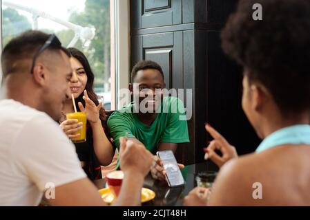 Junger schwarzer Mann, der seinen Freunden etwas auf dem Telefonbildschirm zeigt, während er in einem Café sitzt. Stockfoto