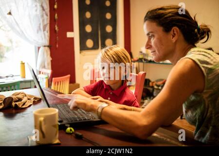 Eine Mutter hilft ihrem Jungen vor einem Laptop, während sie Fernunterricht an einem Tisch zu Hause in Miami, Florida, USA, macht Stockfoto