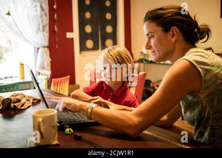 Eine Mutter hilft ihrem kleinen Sohn vor einem Laptop, während sie Fernunterricht an einem Tisch zu Hause in Miami, Florida, USA, macht Stockfoto