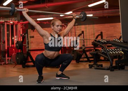 Sportliche junge Frau in Sportkleidung sitzend und Hantelhantel heben Im Fitnessraum Stockfoto