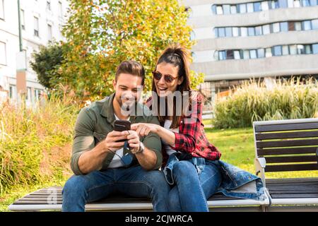 Ein glückliches Paar, das sein Smartphone benutzt und eine App genießt, während es im Sommer auf einer Parkbank in der Stadt sitzt. Stockfoto