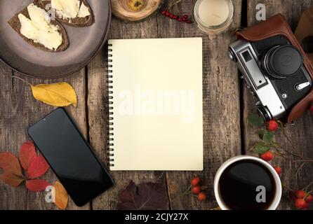 Notizblock, Teller mit Sandwiches, Tasse Kaffee, Kamera, Herbstblätter und Beeren auf natürlichem Holzhintergrund. Stockfoto