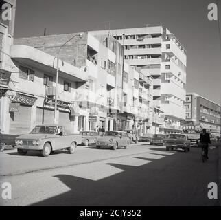 1960er Jahre, Blick aus dieser Ära auf eine staubige, halbgetarnte Straße in Dschidda, Saudi-Arabien, mit Autos aus dieser Zeit und verschiedenen Gebäuden, die am Boden älter und modern sind. Stockfoto