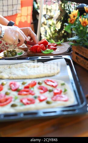 Frau bereitet Focaccia Brot mit geschnittenen ​​tomato, Oliven und frischem Basilikum im Freien. Nahaufnahme. Selektiver Fokus. Italienisches Essen. Stockfoto