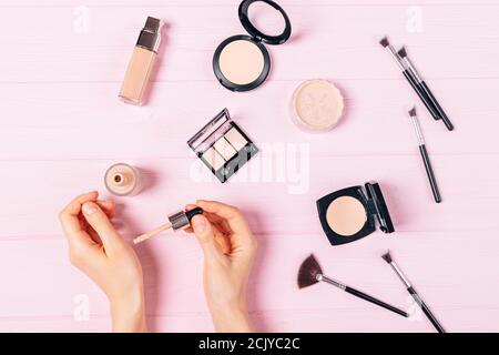 Flach legen Frauen Hände gelten Probe der Stiftung neben Make-up-Produkte und Zubehör für einen gleichmäßigen Teint auf rosa Tisch. Stockfoto