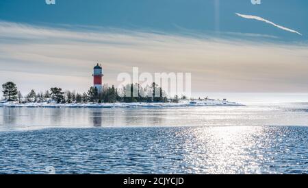 Sehr naher Blick auf den rot-weißen Leuchtturm inmitten einer gefrorenen, verschneiten Insel an der kalten Ostsee, teilweise offenes Wasser, dünnes Eis, das Tageslicht reflektiert. Blau Stockfoto