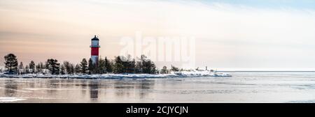 Panorama, Nahaufnahme eines rot-weißen Leuchtturms inmitten einer gefrorenen, verschneiten Insel an der kalten Ostsee, teilweise offenes Wasser, dünnes Eis, das Tageslicht reflektiert. Stockfoto