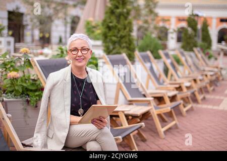 Lächelnd elegante Frau mit einem Rest auf der Straße mit schöner Natur und Gebäude Stockfoto