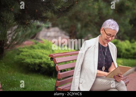 Eine reife Frau mit kurzem Haar wird durch Berühren des Bildschirms des Tablet im Park. Seitenansicht Foto Stockfoto