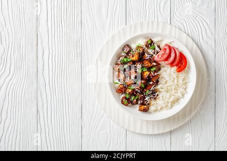 Japanisches Teriyaki gebackenen Tofu mit Reis bestreut von Frühlingszwiebeln, Sesam mit Reis serviert, Tomate in Scheiben geschnitten, auf einer weißen Schüssel auf einem weißen Holz Bac Stockfoto