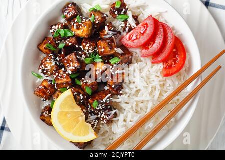 Japanische Teriyaki gebackenen Tofu mit Reis bestreut von Frühlingszwiebeln, Sesam mit Reis serviert, Tomate in Scheiben geschnitten, Zitrone auf einer weißen Schüssel mit Essstäbchen Stockfoto