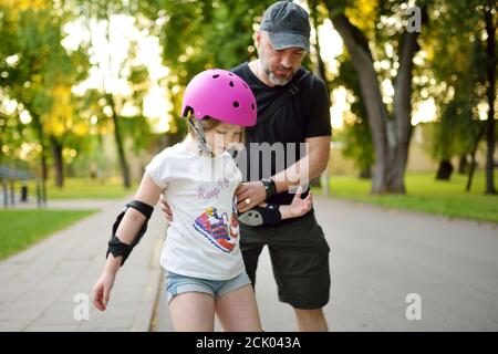 Vater lehrt seine junge Tochter Rollschuh an schönen Sommertag in einem Park. Kind trägt Schutzhelm genießen Rollschuhfahren im Freien Stockfoto