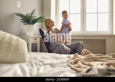Mama mit kleinen Sohn sitzt in einem Stuhl an einem großen Fenster in einem hellen Schlafzimmer. Stockfoto