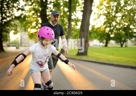 Vater lehrt seine junge Tochter Rollschuh an schönen Sommertag in einem Park. Kind trägt Schutzhelm genießen Rollschuhfahren im Freien Stockfoto