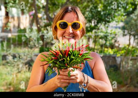 Kaukasische Brünette Frau mit trendigen gelben Sonnenbrille hält Ein Bouquet von frisch geernteten grünen und roten Chilischoten Stockfoto