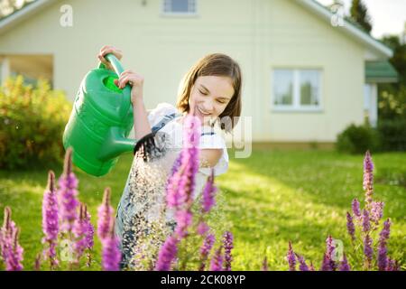 Nettes junges Mädchen, das Blumen im Garten am Sommertag wässert. Kind mit Gießkanne an sonnigen Tag. Mummys kleiner Helfer. Stockfoto