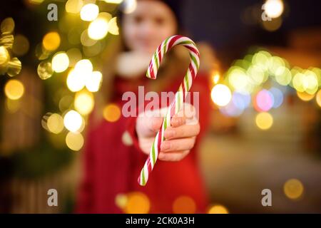 Nettes junges Mädchen mit gestreiften Zuckerrohr auf traditionellen Weihnachtsmarkt in Riga, Lettland. Kinder genießen Süßigkeiten, Bonbons und Lebkuchen auf dem Weihnachtsmarkt Stockfoto