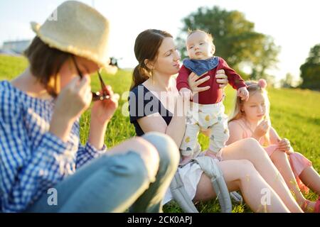 Mutter und drei Kinder, die am Sommertag im Stadtpark Spaß haben. Liebenswert Baby Junge wird von seiner Mama gehalten. Zwei ältere Schwestern umarmen ihre Mutter und BA Stockfoto