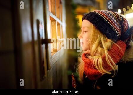 Entzückendes Mädchen, das durch das Fenster auf dem traditionellen Weihnachtsmarkt in Riga, Lettland, guckt. Winterzeit mit Familie und Kindern. Stockfoto