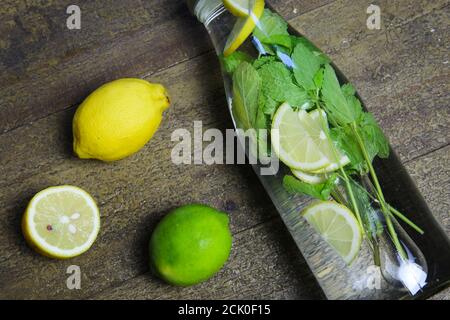 Blick auf isolierte Flasche aus Wasserglas mit Minzblättern und gelben Zitrusfrüchten, grüne Limettenfrüchte auf Holztisch (Fokus auf Flasche) Stockfoto