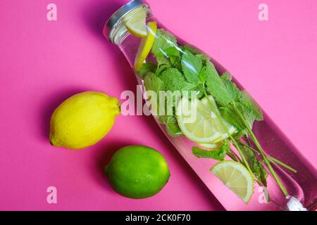 Blick auf isolierte Wasserflasche mit Minzblättern und gelben Zitrusfrüchten, grüne Limettenfrucht auf rosa Hintergrund gewürzt Stockfoto