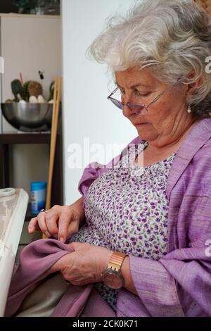 Eine ältere Frau mit grauen Haaren näht bei sich zu Hause von Hand. Sie trägt eine Brille und ein lila Blumenkleid. Stockfoto
