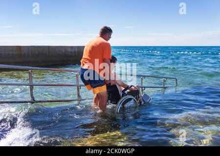 Barrierefreier Strand mit Rampe für Behinderte. Mann im Rollstuhl beim Schwimmen. Stockfoto