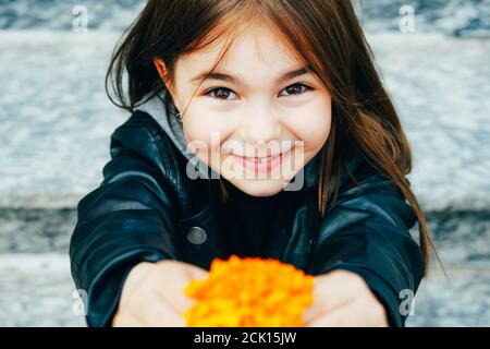 Kleine glückliche Mädchen in stilvollen Herbst-Outfit in einer städtischen Umgebung schaut auf die Kamera und geben eine gelbe Blume. Stockfoto