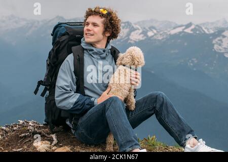 Emotionaler junger Mann mit roten, lockigen Haaren hält seinen Hund und bewundern die Schönheit der Landschaft. Romantischer Ort.blaue Berge auf dem Hintergrund der t Stockfoto