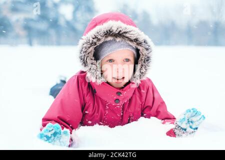 Lustige kaukasische lächelnde Mädchen in warmen Winterkleidung rosa Jacke mit Schnee spielen. Nettes Kind liegt auf dem Boden während kalten Winter schneereichen Tag bei Schneefall Stockfoto