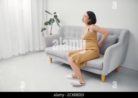 Schöne schwangere Frau, die unter Rückenschmerzen leidet, während sie auf dem Sofa sitzt Während ihrer Schwangerschaft im Wohnzimmer Stockfoto