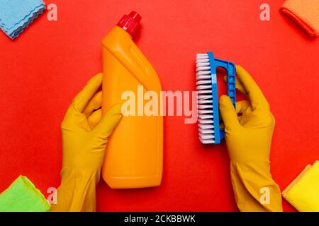 Hände in gelben Gummihandschuhen halten eine gelbe Flasche zum Reinigen der Toilette und eine blaue Bürste auf rotem Hintergrund. Stockfoto