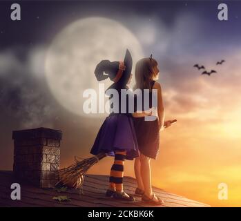 Two happy sisters on Halloween. Funny kids in carnival costumes on the roof with broomstick. Cheerful children looking at moon. Stock Photo