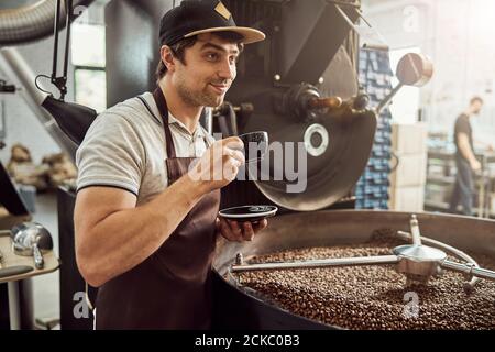Fröhlicher Mann Röster trinkt Kaffee in der Nähe Kaffee Röstermaschine Stockfoto