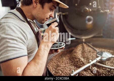 Schöner Mann, der Kaffee in der Nähe der Kaffeeröstermaschine trinkt Stockfoto