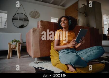 Attraktive afrikanische Frau zu Hause sitzen auf dem Boden mit digitalen Tablet und Kopfhörer blicken weg Stockfoto