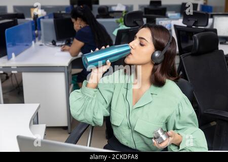 Porträt einer hübschen indischen Geschäftsfrau, die an ihrem Arbeitsplatz sitzt und Wasser aus einer Flasche trinkt, Bürohintergrund, am Laptop arbeitet. Stockfoto