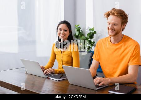 Brunette asiatische Freiberufler mit drahtlosen Kopfhörern auf Hals Blick auf Kamera bei der Verwendung von Laptop in der Nähe Rotschopf Mann Stockfoto