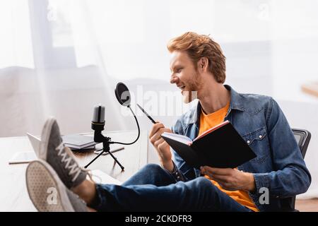 Angeregter Ansager hält Notebook und Stift, während er mit Beinen sitzt Auf dem Schreibtisch und im Mikrofon sprechen Stockfoto