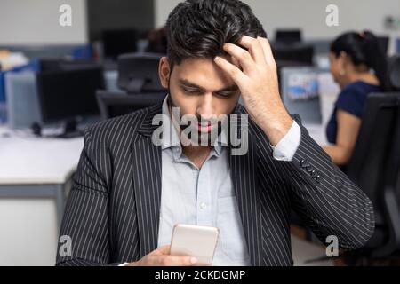 Porträt eines jungen indischen Geschäftsmannes, der vor Bürohintergrund an seinem Arbeitsplatz sitzt, sein Telefon überprüft, seinen Kopf kratzt, Stockfoto