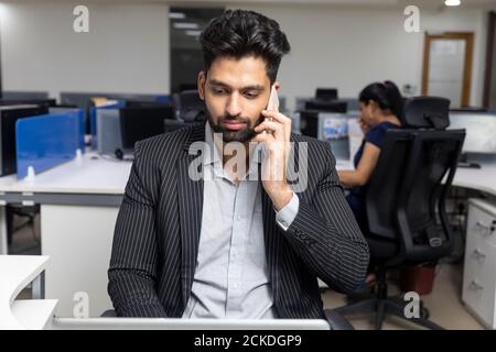 Porträt eines jungen indischen Geschäftsmannes, der am Telefon spricht, an seinem Arbeitsplatz vor Bürohintergrund sitzt, Unternehmensumgebung. Stockfoto