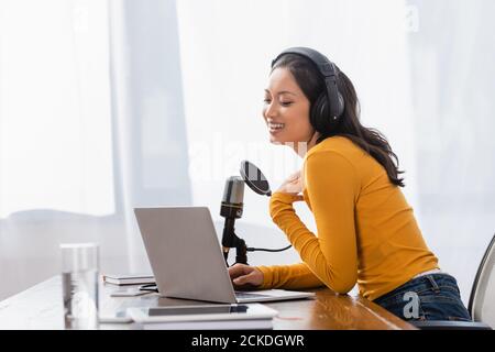 Lachende asiatische Radio-Host in kabellosen Kopfhörern berühren Brust während Arbeiten in der Nähe von Mikrofon und Laptop Stockfoto