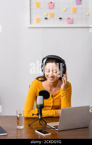 Junge asiatische Radio-Host berühren drahtlose Kopfhörer während sitzen in der Nähe von Mikrofon, Laptop und Smartphone mit leerem Bildschirm Stockfoto