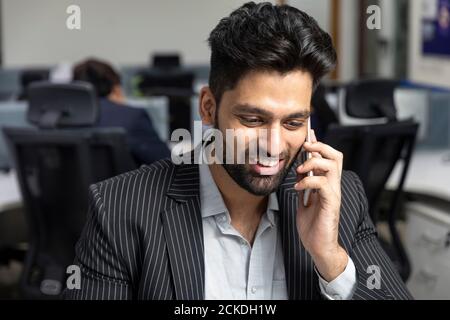 Porträt eines jungen indischen Geschäftsmannes, der am Telefon spricht, an seinem Arbeitsplatz vor Bürohintergrund sitzt, Unternehmensumgebung. Stockfoto
