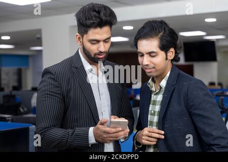 Portrait von zwei jungen gut aussehenden indischen Geschäftsleuten Blick in Telefon, Diskussion Projekt, Unternehmensumfeld. Stockfoto