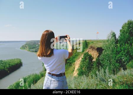 Schöne Frau, die ein Bild von einer Landschaft auf einem Smartphone. Rückansicht, unkenntlich Person.Frau trägt weißes T-Shirt und Jeans fotografieren rote Flagge mit einer Telefonkamera. Reisekonzept. Stockfoto