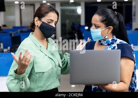 Porträt von zwei jungen indischen Geschäftsfrauen tragen covid Schutzmaske, Blick in Laptop, diskutieren Projekt, Unternehmensumfeld. Stockfoto